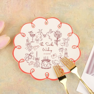 May include: A white, scalloped plate with a red border, decorated with wedding-themed illustrations and the text "The Smiths Wedding 06.12.2026". Two gold forks are placed in front of the plate. The plate's diameter is approximately 20 cm.