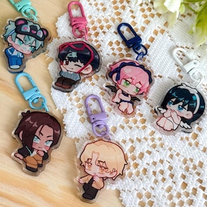 Op de afbeelding: Een verzameling acryl bedels in anime-stijl met kleurrijke karabijnhaaksluitingen. De bedels tonen verschillende personages met unieke kapsels en outfits. De achtergrond omvat wit kant en een houten oppervlak.