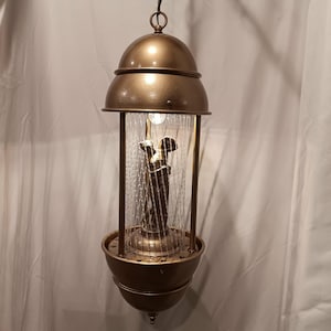 May include: A bronze-coloured hanging lamp with a domed top and a decorative base. The lamp features a central glass enclosure with a sculpture inside, surrounded by a simulated rain effect. The lamp is suspended from a chain.