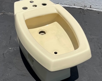 May include: A cream-colored bidet with a rectangular base and an oval basin. The bidet has several holes on the top surface and a central drain. The fixture is made of ceramic or porcelain.
