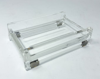 Op de afbeelding: Een heldere, rechthoekige acryl lade met een reeks parallelle ribbels op de basis. De lade heeft een laag profiel met licht verhoogde randen en kleine metalen accenten op elke hoek. Het ontwerp is minimalistisch en functioneel.