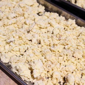 May include: A close-up of a baking sheet filled with a large amount of crumbled, pale yellow dough. The dough is spread evenly across the sheet, creating a textured surface.