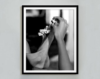 Può includere: Una fotografia in bianco e nero di una persona che si sta dipingendo le unghie dei piedi con un flacone di smalto trasparente. Il piede della persona è appoggiato su una pila di banconote arrotolate.