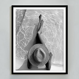 May include: Black and white photograph of a person wearing a wide-brimmed straw hat sitting in a pool of water.