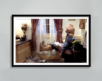 May include: A man in a blue shirt and brown boots sits on a toilet with his legs raised in the air. He is screaming with his mouth open. There are flowers in a vase on the toilet tank.