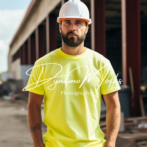 Op de afbeelding: Een man draagt een witte helm met een oranje rand en een felgeel T-shirt met korte mouwen. De man staat voor een gebouw in aanbouw. De tekst "Dynamo Mocks Photography" is zichtbaar op het T-shirt.
