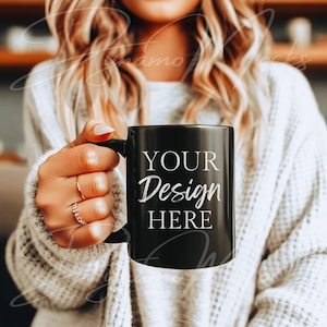 May include: A black coffee mug with the text "YOUR Design HERE" in white script. The mug is held by a person wearing a white sweater. The mug has a black handle and a glossy finish.