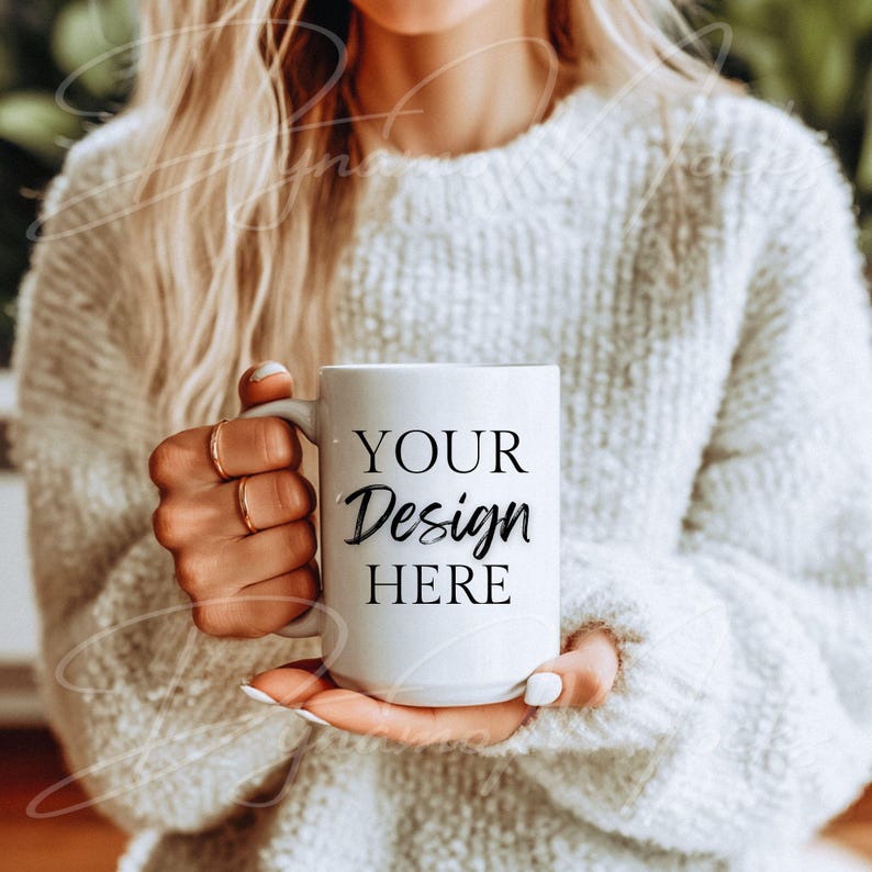 May include: White ceramic mug with the text "YOUR Design HERE" in black script and block letters. The mug is held by a person wearing a white sweater and gold rings. The mug is a standard size.