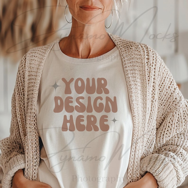 May include: A white t-shirt with the text "YOUR DESIGN HERE" in brown letters with a retro font. The shirt is being worn by a person with a light brown cardigan.