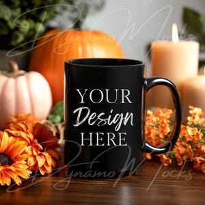 May include: A black coffee mug with the text "YOUR Design HERE" in white. The mug is set against a backdrop of pumpkins, orange flowers, and lit candles, creating a warm, autumnal theme. The mug has a large handle.
