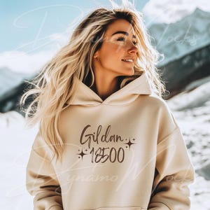 May include: A beige hoodie with the text "Gildan 18500" in brown lettering. The hoodie is worn by a person with long blonde hair, standing in front of a snowy mountain background.