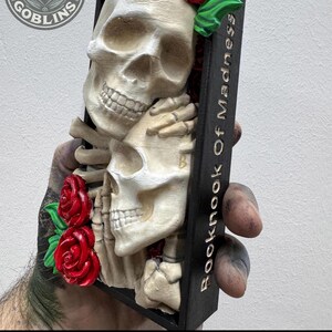 Puede incluir: Una escultura decorativa con dos calaveras abraz&aacute;ndose, adornada con rosas rojas y hojas verdes. La escultura est&aacute; sobre un fondo negro con el texto "Booknook Of Madness" y "Painted by Stacey". El logotipo de Print Goblins es visible.