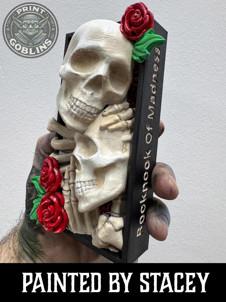 Puede incluir: Una escultura decorativa con dos calaveras abraz&aacute;ndose, adornada con rosas rojas y hojas verdes. La escultura est&aacute; sobre un fondo negro con el texto "Booknook Of Madness" y "Painted by Stacey". El logotipo de Print Goblins es visible.