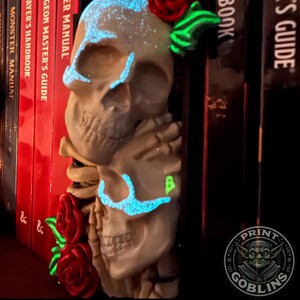 Puede incluir: Una escultura de calaveras apiladas con acentos azules brillantes alrededor de los ojos y la frente, y una rosa roja en la parte superior. La escultura est&aacute; junto a una pila de libros rojos y negros. El texto "PAINTED BY STACEY" est&aacute; en la parte inferior.