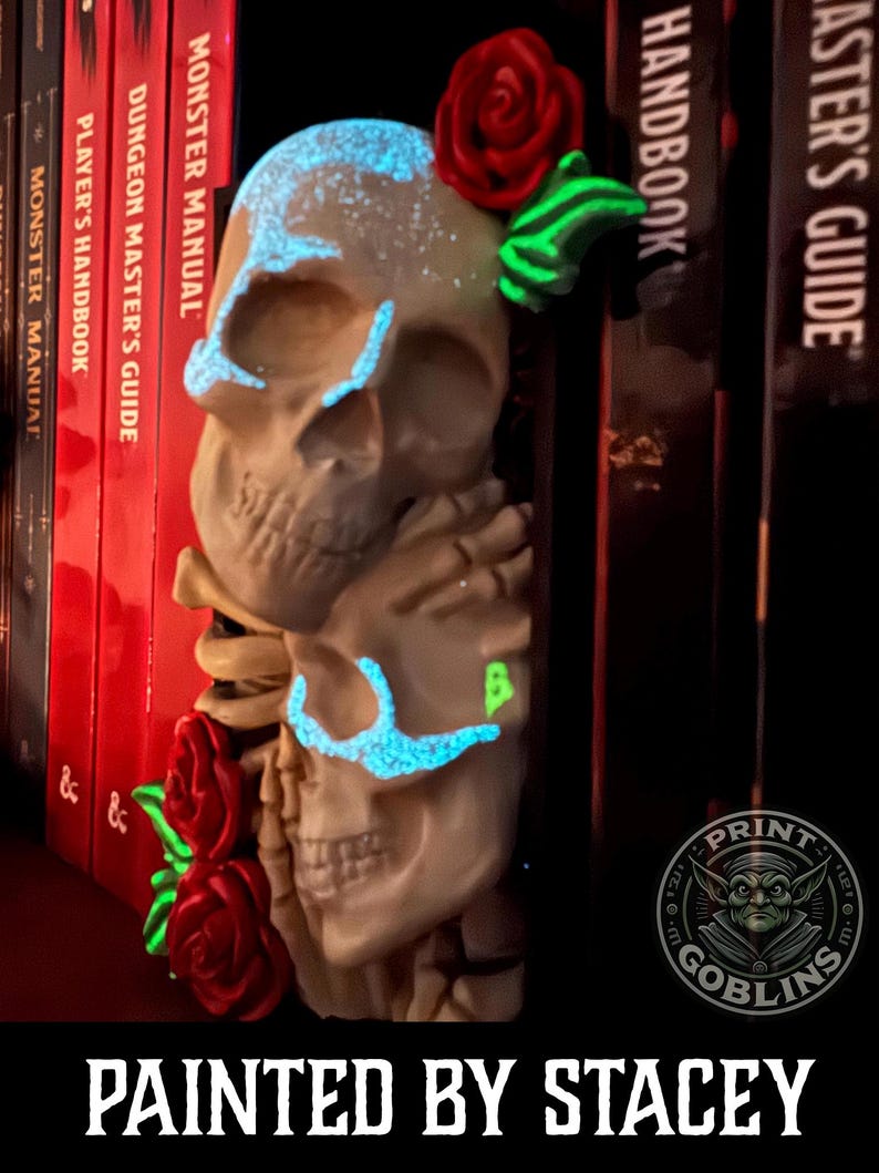 Puede incluir: Una escultura de calaveras apiladas con acentos azules brillantes alrededor de los ojos y la frente, y una rosa roja en la parte superior. La escultura est&aacute; junto a una pila de libros rojos y negros. El texto "PAINTED BY STACEY" est&aacute; en la parte inferior.