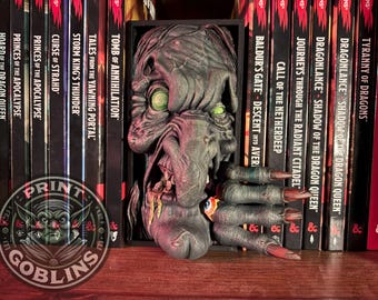 May include: A detailed, gray monster bookend with green eyes and long, clawed fingers. The bookend is placed between a row of fantasy books with titles like "Tomb of Annihilation" and "Tyranny of Dragons". A circular logo with the text "Print Goblins" is visible.
