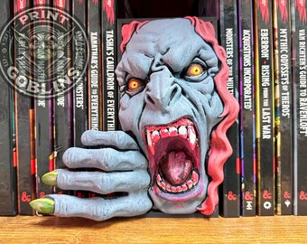 Op de afbeelding: Een grijs en rood monster-thema boekensteun met een gedetailleerd gezicht en handontwerp. De boekensteun staat tussen fantasyboeken, wat een uniek decoratief element aan een boekenplank toevoegt.