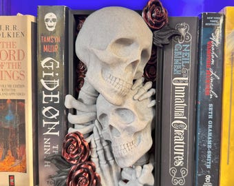 Puede incluir: Un sujetalibros gris con forma de calavera con dos calaveras abrazándose, rodeado de rosas rojas. El sujetalibros se coloca entre varios libros, incluyendo "Gideon the Ninth" y "Unnatural Creatures". Un artículo decorativo único.