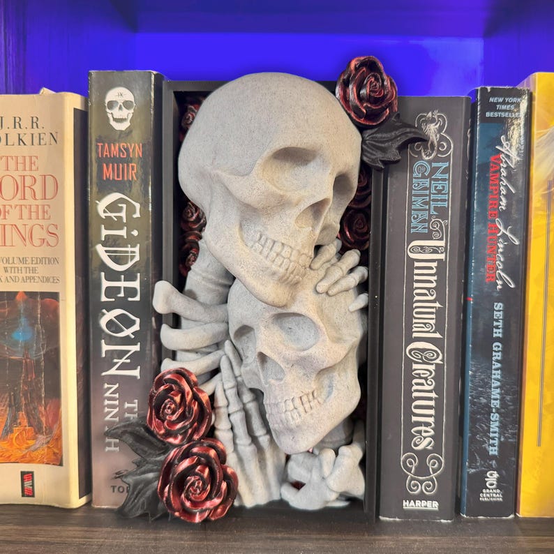 Puede incluir: Un sujetalibros gris con forma de calavera con dos calaveras abraz&aacute;ndose, rodeado de rosas rojas. El sujetalibros se coloca entre varios libros, incluyendo "Gideon the Ninth" y "Unnatural Creatures". Un art&iacute;culo decorativo &uacute;nico.