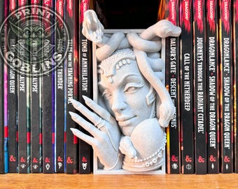 May include: A detailed, gray Medusa bookend sculpture. The bookend features Medusa's face, snakes, and a hand with rings. The bookend is placed between several fantasy books with titles like "Tomb of Annihilation" and "Dragonlance".