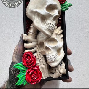 Puede incluir: Escultura detallada de calaveras en blanco roto con rosas rojas y hojas verdes, enmarcada en negro. El texto "PAINTED BY STACEY" est&aacute; en la parte inferior. La obra representa dos calaveras entrelazadas.