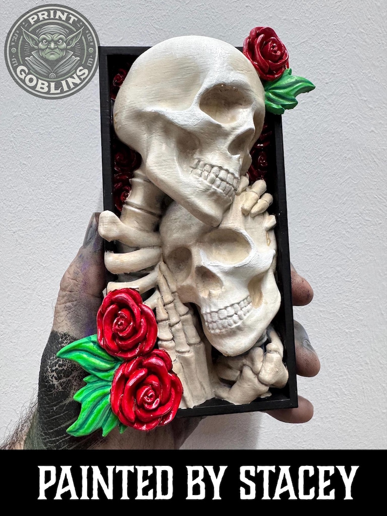 Puede incluir: Escultura detallada de calaveras en blanco roto con rosas rojas y hojas verdes, enmarcada en negro. El texto "PAINTED BY STACEY" est&aacute; en la parte inferior. La obra representa dos calaveras entrelazadas.