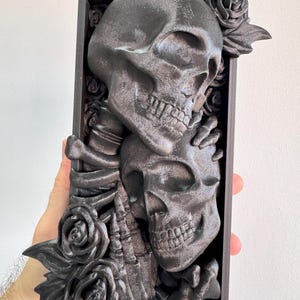 Puede incluir: Una pieza de arte mural rectangular gris oscuro con dos calaveras abraz&aacute;ndose, rodeadas de rosas. Las calaveras est&aacute;n intrincadamente detalladas, con dientes y cuencas de los ojos visibles. Las rosas tambi&eacute;n est&aacute;n esculpidas, a&ntilde;adiendo una est&eacute;tica g&oacute;tica y rom&aacute;ntica.