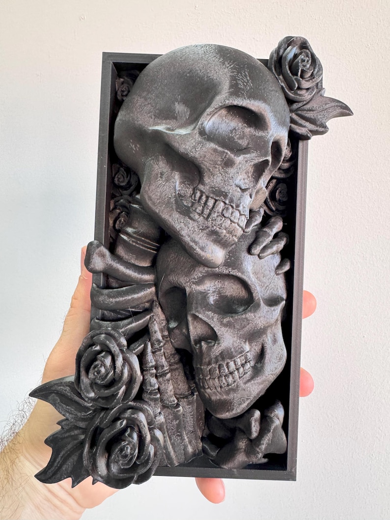 Puede incluir: Una pieza de arte mural rectangular gris oscuro con dos calaveras abraz&aacute;ndose, rodeadas de rosas. Las calaveras est&aacute;n intrincadamente detalladas, con dientes y cuencas de los ojos visibles. Las rosas tambi&eacute;n est&aacute;n esculpidas, a&ntilde;adiendo una est&eacute;tica g&oacute;tica y rom&aacute;ntica.
