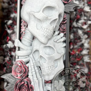 Puede incluir: Una escultura gris que presenta dos calaveras entrelazadas, acentuadas con rosas rojas y negras. Las calaveras est&aacute;n dentro de un marco rectangular. El logotipo de Print Goblins es visible en la esquina inferior derecha.