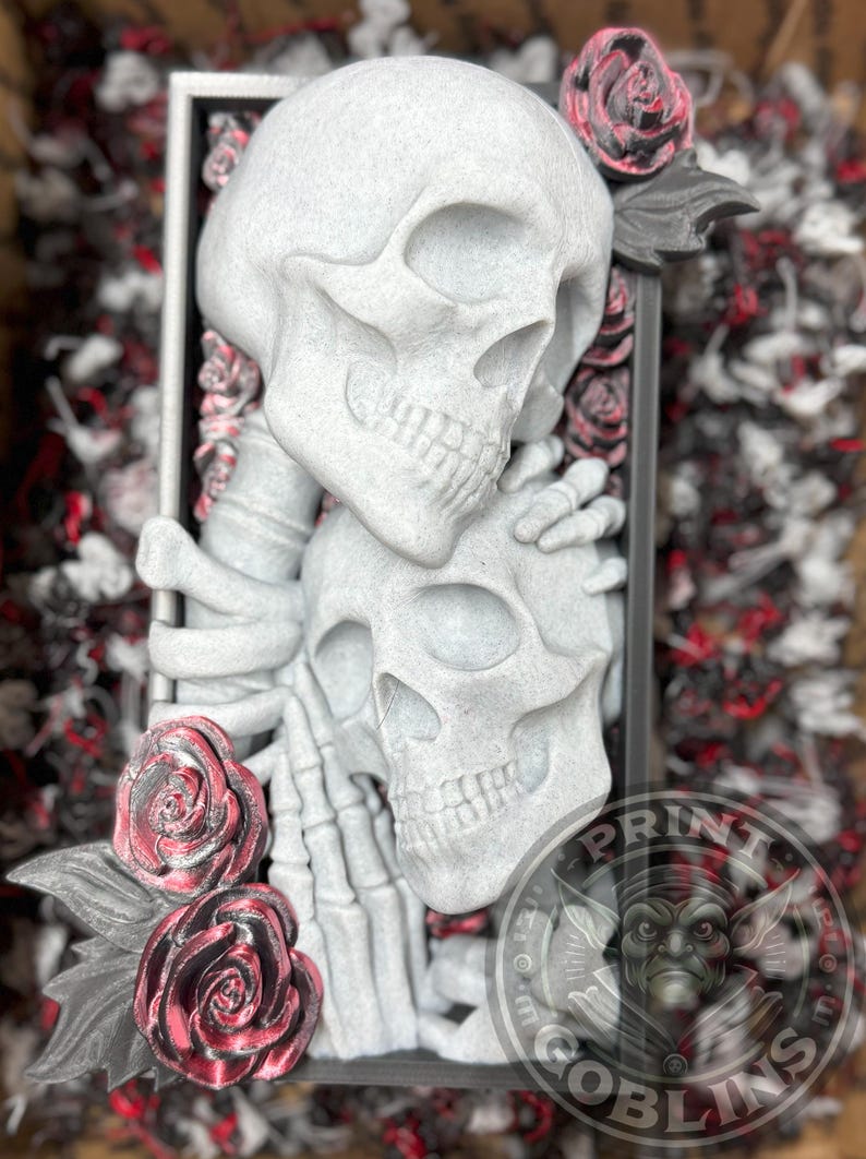 Puede incluir: Una escultura gris que presenta dos calaveras entrelazadas, acentuadas con rosas rojas y negras. Las calaveras est&aacute;n dentro de un marco rectangular. El logotipo de Print Goblins es visible en la esquina inferior derecha.