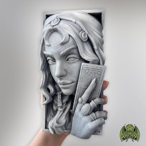 May include: A rectangular gray sculpture featuring a relief of a face with wavy hair and a headband. A hand with rings is positioned in the foreground, holding a rectangular card with etched line details. The entire piece has a textured finish.