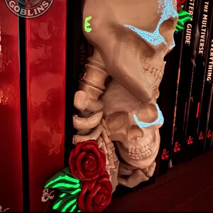 Puede incluir: Una decoraci&oacute;n de estanter&iacute;a con forma de calavera beige con detalles azules brillantes y rosas rojas. El texto "PAINTED BY STACEY" est&aacute; en la parte inferior. El logotipo de Print Goblins est&aacute; en la esquina superior izquierda. Decoraci&oacute;n frente a libros rojos.