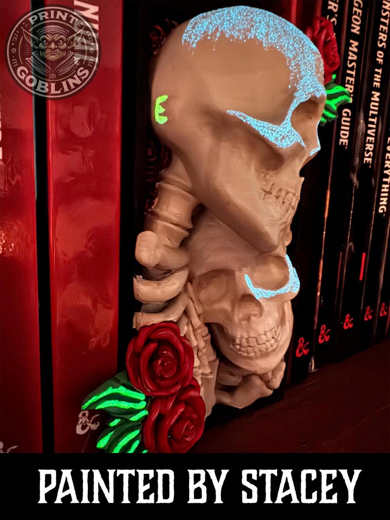 Puede incluir: Una decoraci&oacute;n de estanter&iacute;a con forma de calavera beige con detalles azules brillantes y rosas rojas. El texto "PAINTED BY STACEY" est&aacute; en la parte inferior. El logotipo de Print Goblins est&aacute; en la esquina superior izquierda. Decoraci&oacute;n frente a libros rojos.