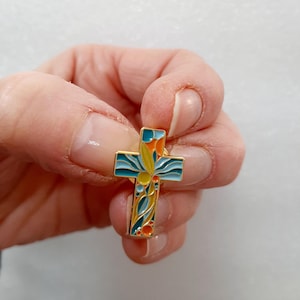 Peut inclure: Une épingle en émail dorée en forme de croix. La croix est décorée d'un motif floral coloré dans les tons bleu, vert, jaune et orange.