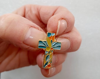 Puede incluir: Un pin de esmalte dorado en forma de cruz. La cruz está decorada con un diseño floral colorido en tonos azules, verdes, amarillos y naranjas.