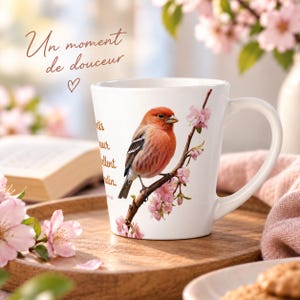 Peut inclure: Un mug en céramique blanche orné d'une illustration détaillée d'un oiseau à poitrine rouge perché sur une branche fleurie aux pétales roses. Le mug porte le texte "Un moment de douceur" ainsi que du texte manuscrit partiel.