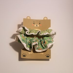 May include: Green and white floral fabric scrunchie with a white lace trim. The scrunchie is displayed on a brown cardboard bear-shaped card with the word "Scrunchies" printed on it.