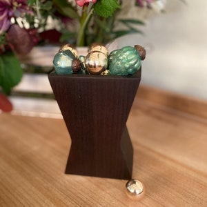 Peut inclure: Un vase en bois brun foncé avec une forme de sablier unique, rempli d'une collection d'ornements en verre vert et or.