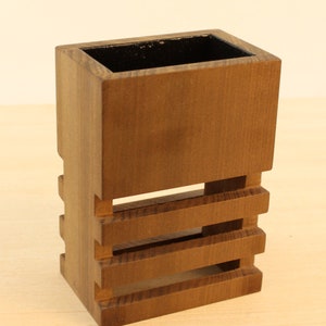 May include: A wooden pencil holder with a dark brown stain. The holder has three open slats on the front and sides.