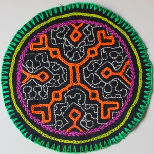 Pode incluir: Um emblema circular de tecido preto com bordado manual intrincado. O design inclui um padrão geométrico laranja central rodeado por linhas cinza-prateadas, uma borda circular magenta e uma borda externa costurada em ziguezague verde brilhante.