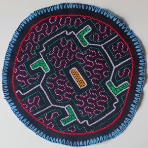 Pode incluir: Um emblema circular de tecido preto com bordado geométrico intrincado em fios rosa, verde, roxo claro e amarelo. A borda é finalizada com um ponto caseado azul claro e uma fina linha circular vermelha emoldura o padrão central.