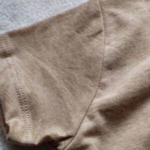 May include: Close-up of a tan short-sleeve shirt. The sleeve's edge is visible, showcasing a neatly stitched hem. The fabric has a textured appearance, with subtle variations in color. The shirt appears to be made of a soft, breathable material.