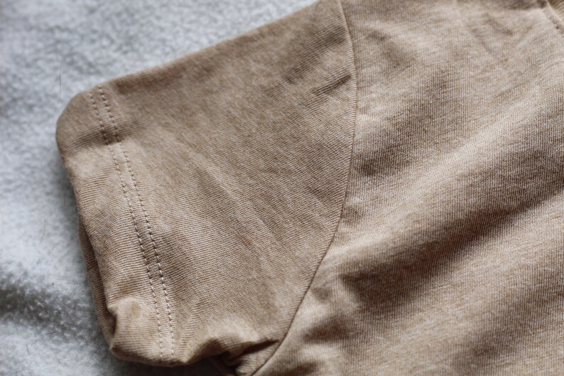 May include: Close-up of a tan short-sleeve shirt. The sleeve's edge is visible, showcasing a neatly stitched hem. The fabric has a textured appearance, with subtle variations in color. The shirt appears to be made of a soft, breathable material.