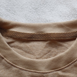 May include: Close-up of a tan-colored crew neck t-shirt. The neckline is ribbed with a brown herringbone tape detail on the inside. The shirt is made of a soft, comfortable material, perfect for casual wear.