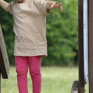 May include: A young child wearing a tan t-shirt and pink leggings is balancing on a wooden structure in a park. The child has their arms outstretched for balance. The wooden structure has brown and gray elements.