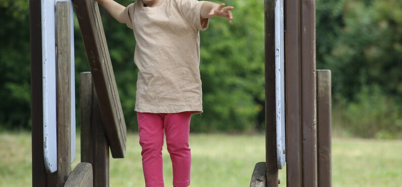 May include: A young child wearing a tan t-shirt and pink leggings is balancing on a wooden structure in a park. The child has their arms outstretched for balance. The wooden structure has brown and gray elements.