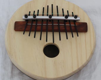 May include: A round wooden kalimba with a light wood finish. It features a dark brown soundboard and a series of black metal tines. The instrument has a circular sound hole and a metal bridge.