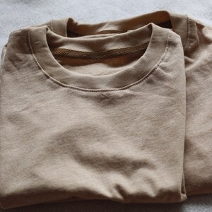 May include: Two folded, tan-colored crew neck t-shirts. The shirts are made of a soft material and are neatly stacked on top of each other. The collars have a ribbed texture.