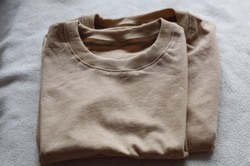 May include: Two folded, tan-colored crew neck t-shirts. The shirts are made of a soft material and are neatly stacked on top of each other. The collars have a ribbed texture.