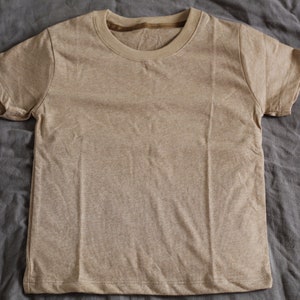 May include: A short-sleeved, tan-colored t-shirt. The shirt has a crew neck and is made of a soft, textured fabric. The t-shirt is laying flat on a gray, wrinkled surface.